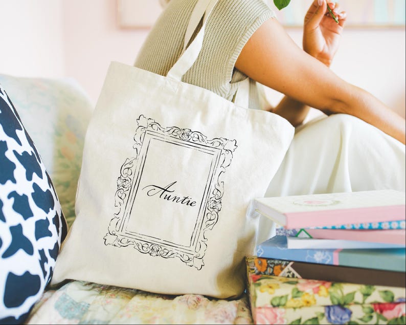 May include: Cream-colored tote bag with a black ornate frame design and the word "Auntie" printed inside. The bag has a simple strap and is sitting next to a stack of books with floral and pastel covers.