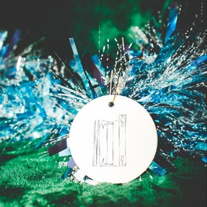 May include: A round, white ornament with a sketched illustration of three books. The ornament hangs from a gold string against a backdrop of blue and silver tinsel and a green, textured surface.