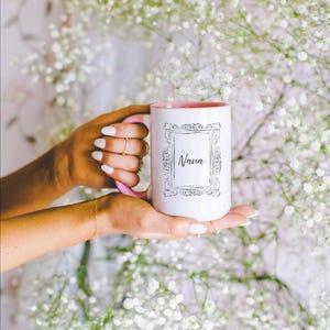 May include: White ceramic mug with a pink interior and handle, featuring the word "Nana" in a decorative frame. The mug is held against a backdrop of white flowers.