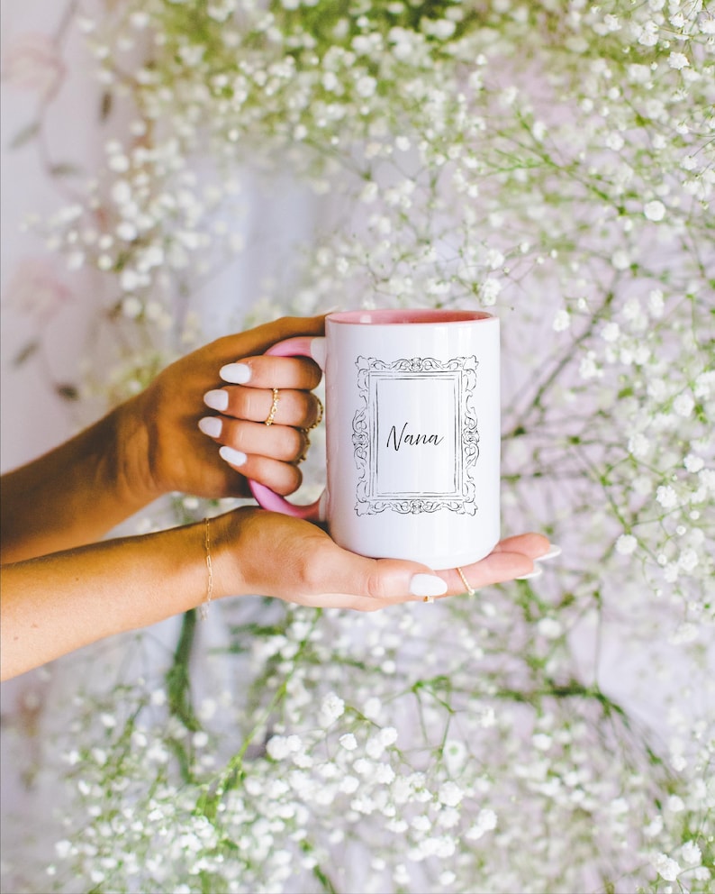 May include: White ceramic mug with a pink interior and handle, featuring the word "Nana" in a decorative frame. The mug is held against a backdrop of white flowers.