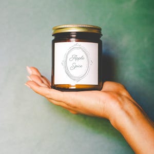 May include: A brown glass jar with a gold lid, labeled "Apple Spice" in an oval frame. The jar is held in a person's hand against a teal background. The label is white with black text.
