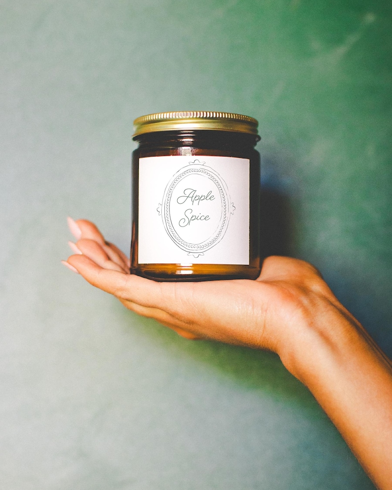 May include: A brown glass jar with a gold lid, labeled "Apple Spice" in an oval frame. The jar is held in a person's hand against a teal background. The label is white with black text.
