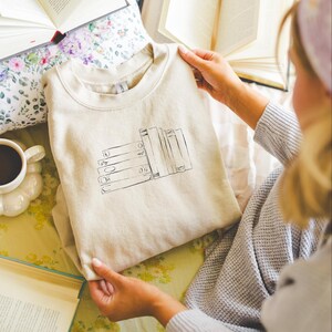 May include: Beige sweatshirt with a black graphic of stacked books. The sweatshirt is folded and held by a person. Open books and a cup of coffee are in the background, suggesting a reading theme.