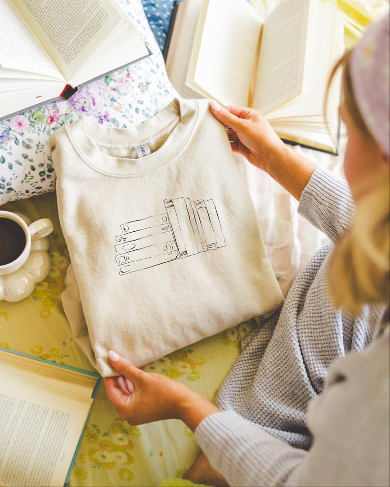 May include: Beige sweatshirt with a black graphic of stacked books. The sweatshirt is folded and held by a person. Open books and a cup of coffee are in the background, suggesting a reading theme.
