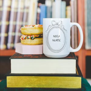 May include: A white ceramic mug with the text "NICU NURSE" inside a decorative oval frame, topped with a bow. The mug sits atop a stack of books, next to two donuts with colorful sprinkles. The background features a bookshelf.