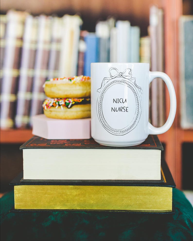 May include: A white ceramic mug with the text "NICU NURSE" inside a decorative oval frame, topped with a bow. The mug sits atop a stack of books, next to two donuts with colorful sprinkles. The background features a bookshelf.