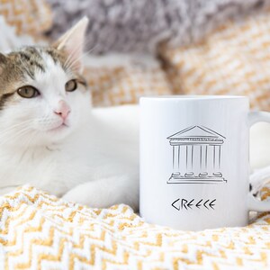 May include: White ceramic mug with a black line drawing of a Greek temple and the word "GREECE" written in Greek letters. A white cat is lying on a yellow and white blanket behind the mug.