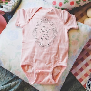 May include: A light pink baby onesie with the text "the fairest one of all" inside a decorative frame. The onesie is short-sleeved and has a snap closure. The background includes pillows and disco balls.