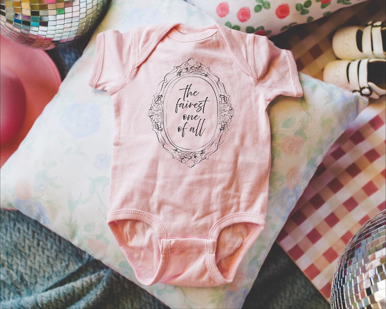 May include: A light pink baby onesie with the text "the fairest one of all" inside a decorative frame. The onesie is short-sleeved and has a snap closure. The background includes pillows and disco balls.