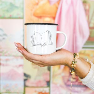 May include: White enamel mug with a black rim featuring a line drawing of an open book. The mug is held in a hand, with a floral bracelet visible on the wrist. The background includes colorful artwork and a pink bow.