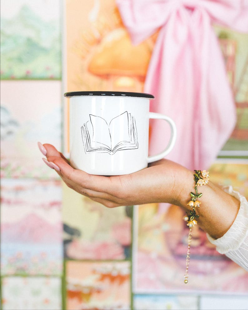 May include: White enamel mug with a black rim featuring a line drawing of an open book. The mug is held in a hand, with a floral bracelet visible on the wrist. The background includes colorful artwork and a pink bow.
