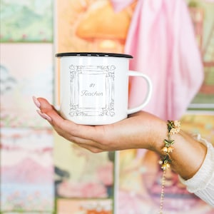 May include: White enamel mug with a black rim, featuring the text "#1 Teacher" inside a decorative frame. The mug is held in a hand, with a floral bracelet visible on the wrist. The background includes colorful artwork and a pink bow.