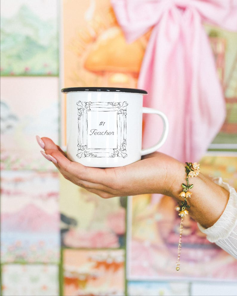 May include: White enamel mug with a black rim, featuring the text "#1 Teacher" inside a decorative frame. The mug is held in a hand, with a floral bracelet visible on the wrist. The background includes colorful artwork and a pink bow.