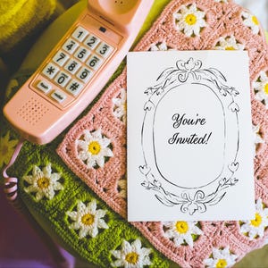 May include: A vintage-style scene featuring a pink rotary phone and a greeting card that says "You're Invited!" The card is set on a handmade crochet blanket with daisy patterns in pink, green, and yellow. The phone is a retro design.