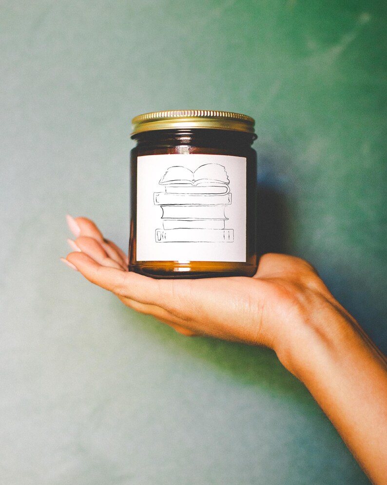 May include: A brown glass jar with a gold lid, featuring a white label with a line drawing of stacked books. The jar is held in a hand against a green background. The jar appears to be a candle.