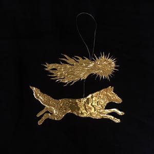 Op de afbeelding: Een goudkleurig metalen ornament met een rennende wolf onder een gestileerde komeet- of zonnestraalvorm. Het ornament hangt aan een zilveren draad tegen een zwarte achtergrond. De wolf is gedetailleerd met textuurpatronen.