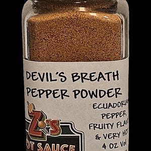 May include: A clear glass jar filled with brown pepper powder. The label reads "Devil's Breath Pepper Powder" and includes text about the pepper's origin and flavor profile. The jar has a black lid and a label from Z's Hot Sauce.