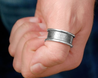 Könnte beinhalten: Ein silberner Ring mit einem breiten Band und einer strukturierten Oberfläche. Der Ring wird am Daumen getragen.