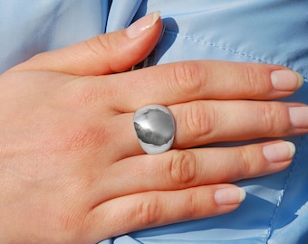 May include: A silver ring with a large, smooth, oval-shaped top. The ring is worn on the middle finger of a hand.