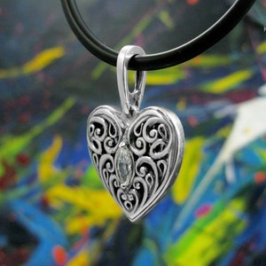 May include: A silver heart-shaped pendant with an intricate filigree design and a clear gemstone in the center. The pendant is hanging from a black leather cord.
