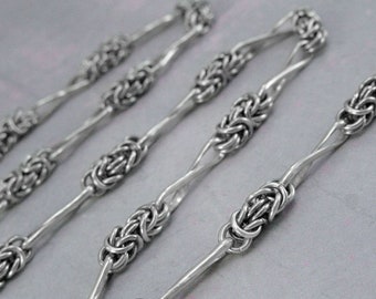 May include: A silver chain necklace with a unique, intricate design. The chain is made of small, linked rings that create a textured, woven look.