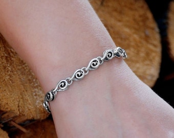 May include: A silver bracelet with a repeating swirl design is worn on a wrist. The bracelet is made of linked metal pieces. The background is a stack of brown logs.