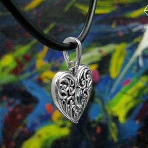 May include: A silver heart-shaped pendant with intricate scrollwork and a clear crystal in the center. The pendant is hanging from a black leather cord.