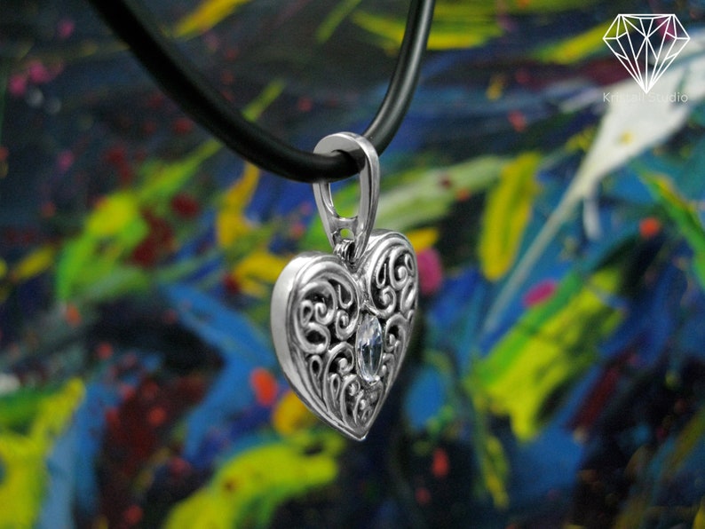 May include: A silver heart-shaped pendant with intricate scrollwork and a clear crystal in the center. The pendant is hanging from a black leather cord.