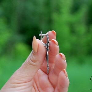 May include: Silver dangle earrings with a floral design. The earrings are held in a hand with a green background.