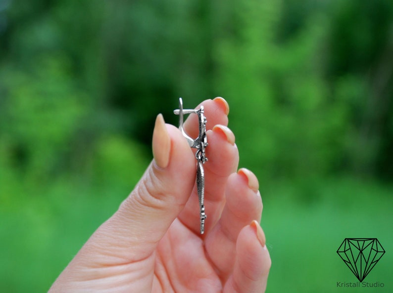 May include: Silver dangle earrings with a floral design. The earrings are held in a hand with a green background.