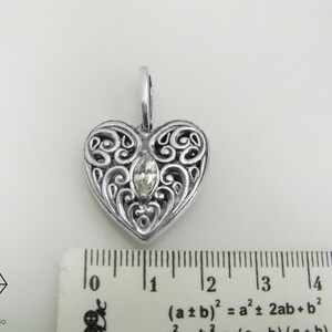May include: A silver heart-shaped pendant with an intricate filigree design and a clear gemstone in the center. The pendant measures approximately 1 inch in length.