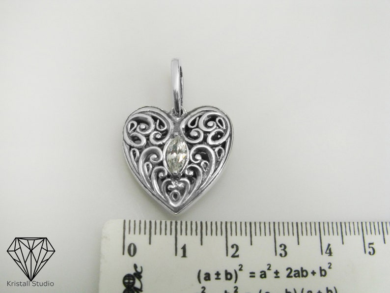 May include: A silver heart-shaped pendant with an intricate filigree design and a clear gemstone in the center. The pendant measures approximately 1 inch in length.