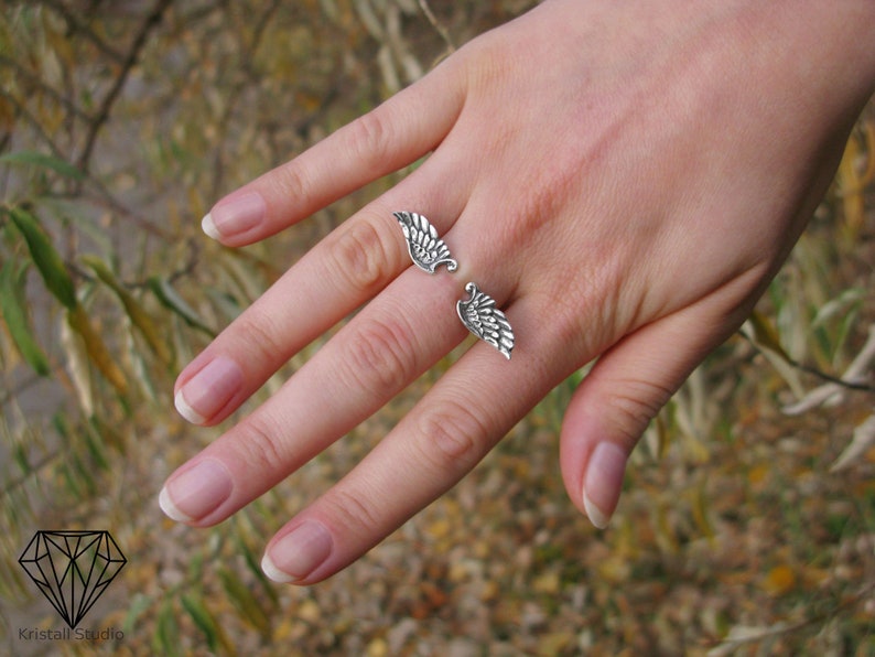 May include: A silver ring with two wings on the band. The ring is worn on the middle finger of a hand.