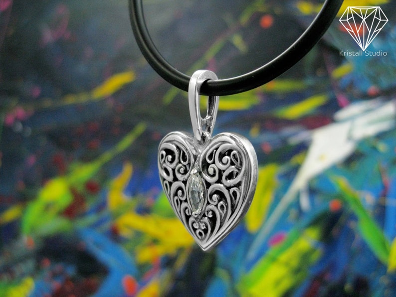 May include: A silver heart-shaped pendant with an intricate filigree design and a clear gemstone in the center. The pendant is hanging from a black leather cord.