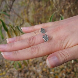 May include: A silver ring with two wings on the band. The ring is on a person's finger.