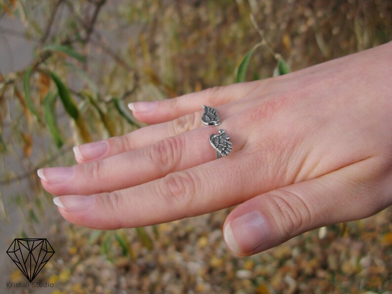 May include: A silver ring with two wings on the band. The ring is on a person's finger.