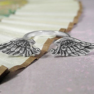 May include: A silver ring with two angel wings on either side. The wings are detailed and have a textured finish.