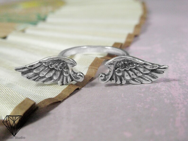 May include: A silver ring with two angel wings on either side. The wings are detailed and have a textured finish.