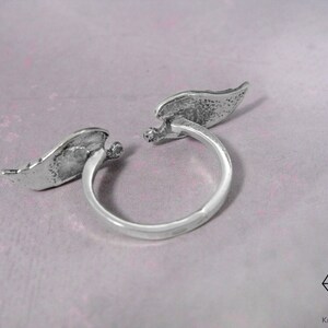 May include: Silver ring with two angel wings on either side of the band. The wings are textured and have a slightly distressed look.