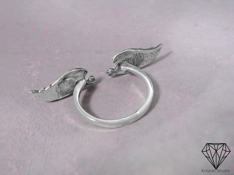 May include: Silver ring with two angel wings on either side of the band. The wings are textured and have a slightly distressed look.