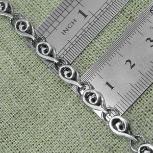 May include: Close-up of a silver chain necklace with a repeating heart-shaped design. The chain is measured against a ruler, showing it is approximately 1 inch long.