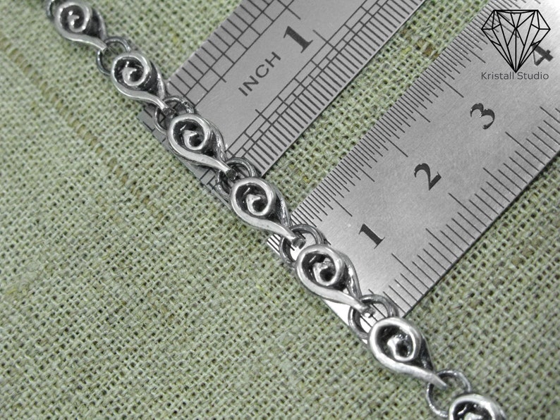 May include: Close-up of a silver chain necklace with a repeating heart-shaped design. The chain is measured against a ruler, showing it is approximately 1 inch long.