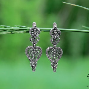 May include: A pair of silver filigree earrings with a heart-shaped design. The earrings are hanging from a green stem.