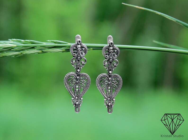May include: A pair of silver filigree earrings with a heart-shaped design. The earrings are hanging from a green stem.
