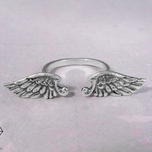 May include: A silver ring with two angel wings on either side of the band.