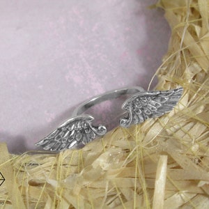 May include: A silver ring with two angel wings on the band. The ring is on a light brown background.