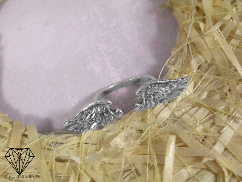 May include: A silver ring with two angel wings on the band. The ring is on a light brown background.