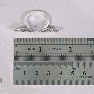May include: A silver adjustable ring with two angel wings on either side. The ring is measured against a ruler with inch and centimeter markings.