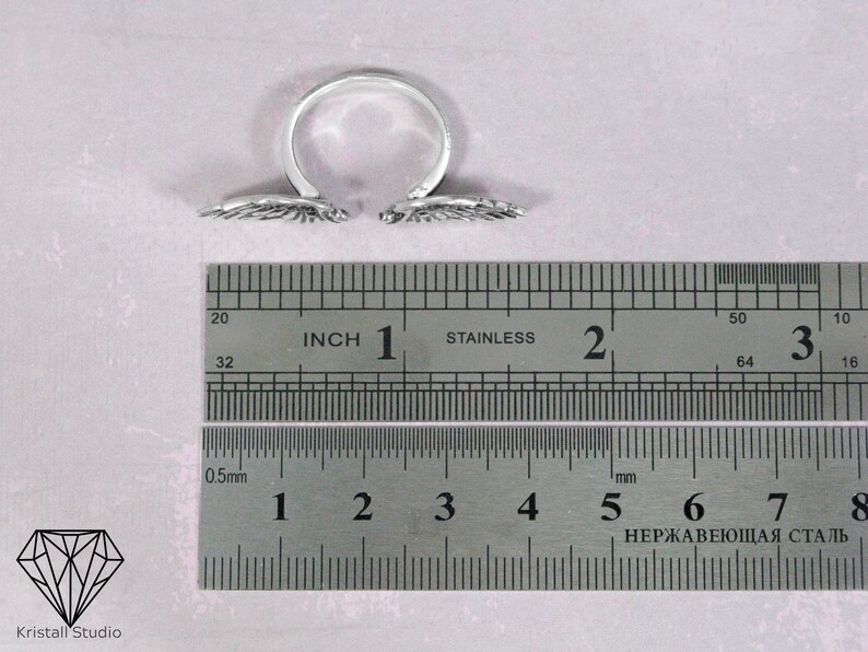 May include: A silver adjustable ring with two angel wings on either side. The ring is measured against a ruler with inch and centimeter markings.