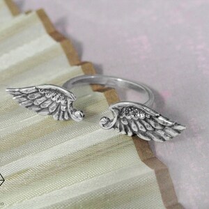 May include: A silver ring with two angel wings on either side. The wings are detailed and have a textured finish.